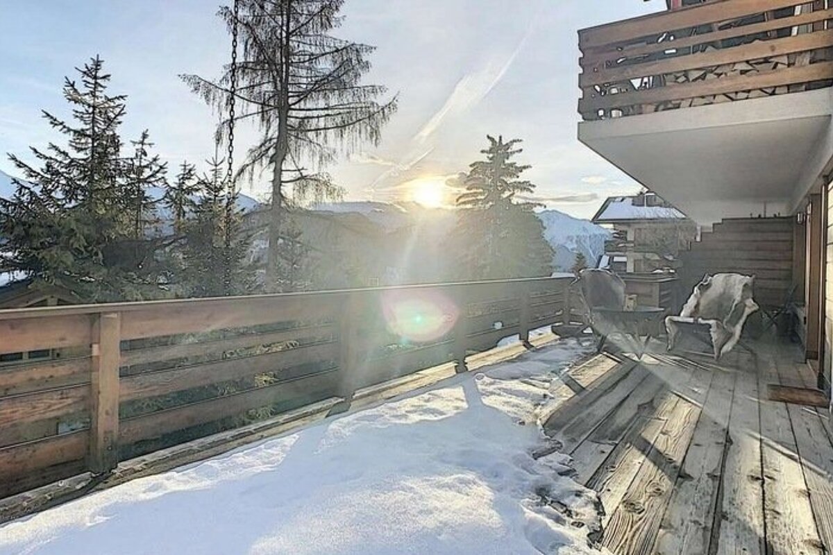 Snowy wooden balcony of an alpine chalet overlooking pine trees and distant mountains under a bright sun.
