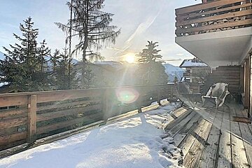Snowy wooden balcony of an alpine chalet overlooking pine trees and distant mountains under a bright sun.
