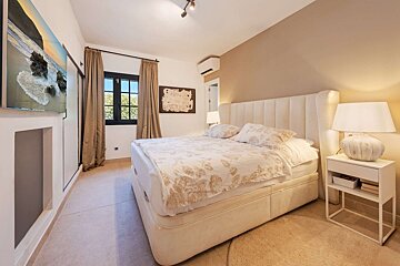 A bright, neutral-toned bedroom with a large bed, window, TV featuring a beach, nightstands, and lamps, creating a cozy and modern feel.