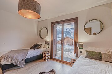 Cozy bedroom with two twin beds, large wooden-framed windows opening to a snowy mountain view and a nearby cabin. Modern decor.