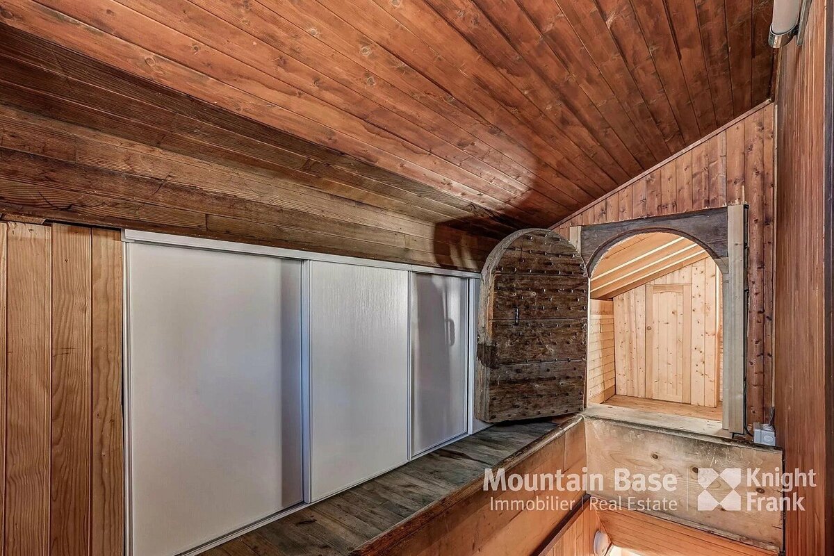 A rustic wood-paneled attic with a slanted ceiling, built-in white sliding storage, and an arched wooden door open to another timber room.