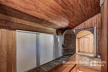 A cozy, rustic wooden attic with a sloped ceiling, white sliding doors, and an arched opening to another wood-paneled room. A unique wooden door is open.