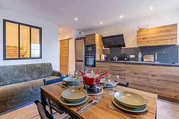 Modern open-plan living/dining/kitchen. A wooden table is set with a fondue pot, next to an olive green sofa and contemporary kitchen.