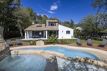 A charming white house with a tiled roof sits beside a unique rock-edged pool and spa, surrounded by lush greenery under a clear blue sky.