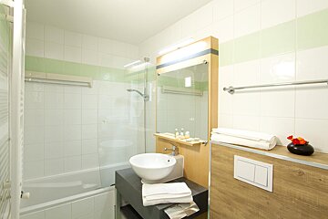 A bright bathroom with white tiles and a green stripe, featuring a bathtub with shower, bowl sink, large mirror, and neatly folded towels.