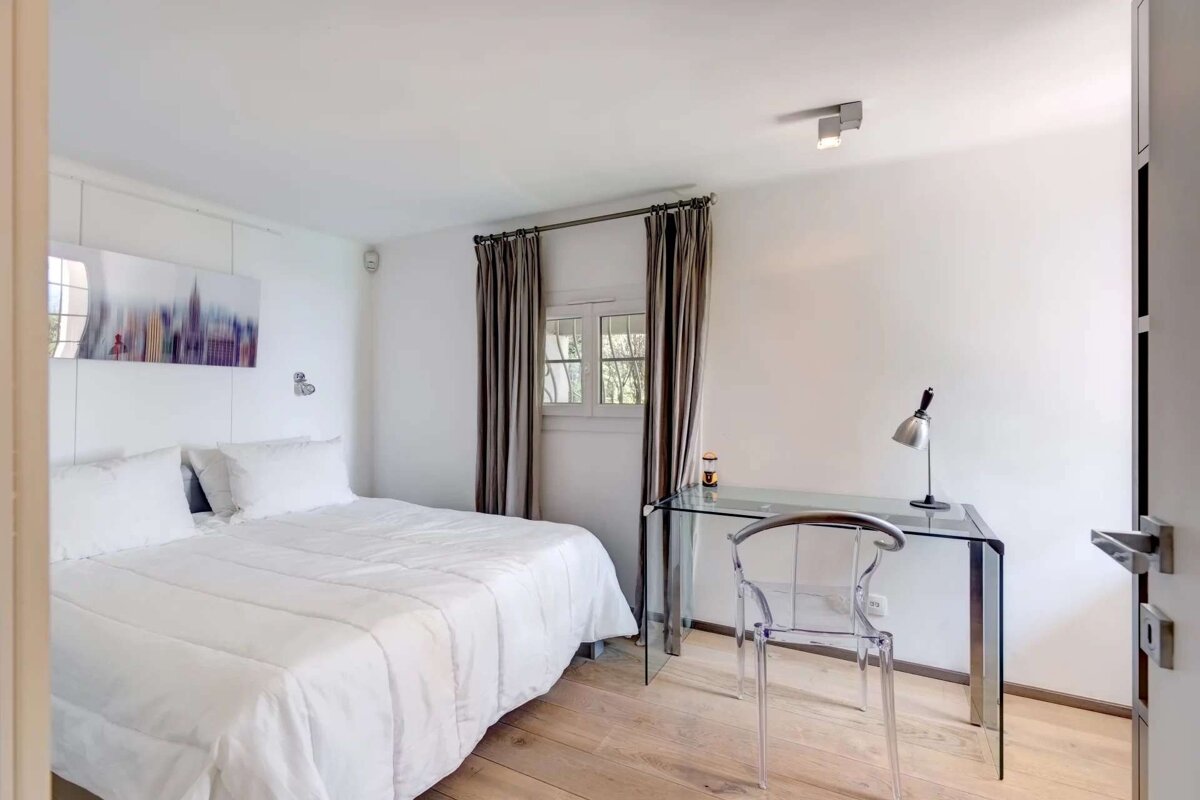 Bright, minimalist bedroom featuring a bed with white linen, wooden floor, glass desk with clear chair, and window with taupe curtains.