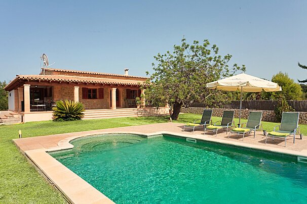 Sunny stone villa with a tiled roof, a clear turquoise swimming pool, green lawn, sun loungers, and an umbrella under a bright blue sky.