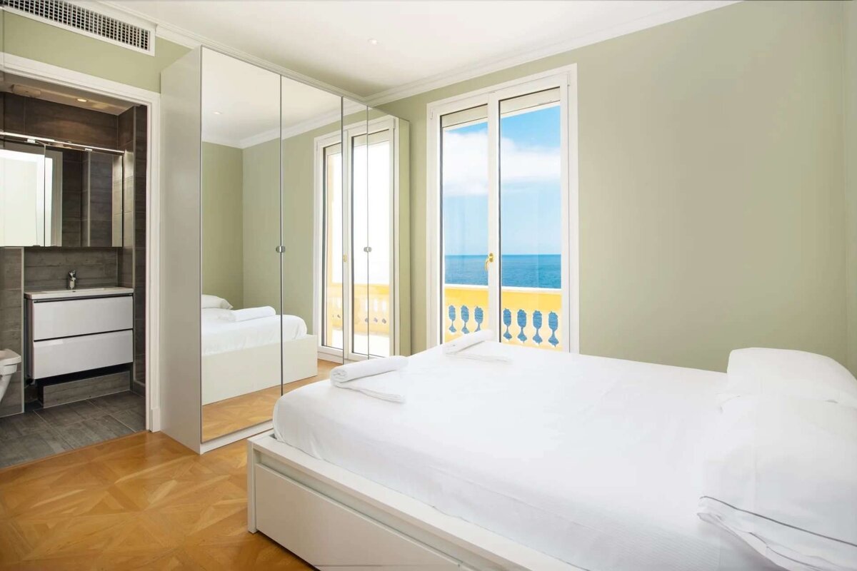 A bright bedroom with a white bed, mirrored wardrobe, en-suite bathroom, and a balcony offering sea views. Pale green walls.