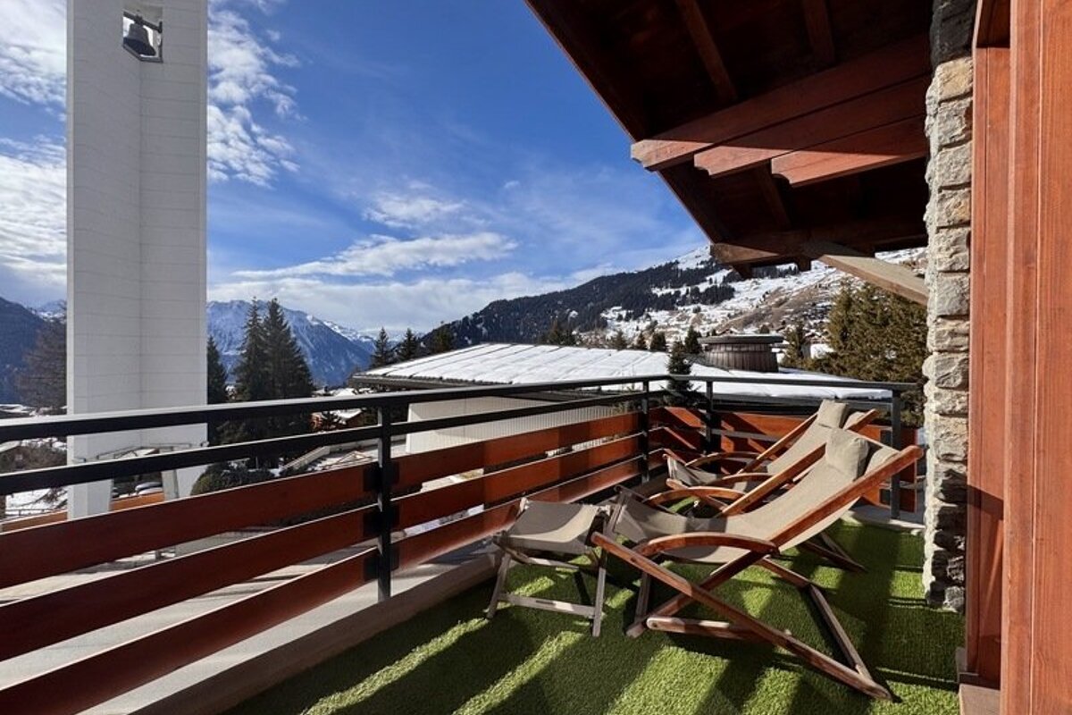 Wooden lounge chairs on a chalet balcony overlooking snowy mountains and a white bell tower under a blue sky.