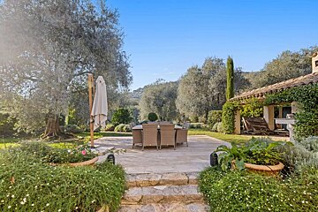 A sunny outdoor dining patio with wicker furniture, surrounded by olive trees, lush greenery, and a tiled-roof building under a clear blue sky.