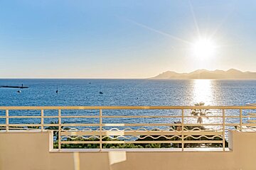 Sunlit ocean view from a balcony with white railings, featuring boats on the water and mountains in the distance.