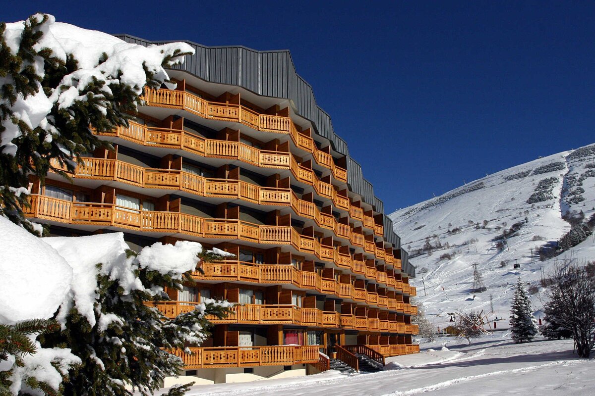 A multi-story ski resort building with numerous wooden balconies is covered in snow, set against a clear blue sky and snowy mountain slopes.
