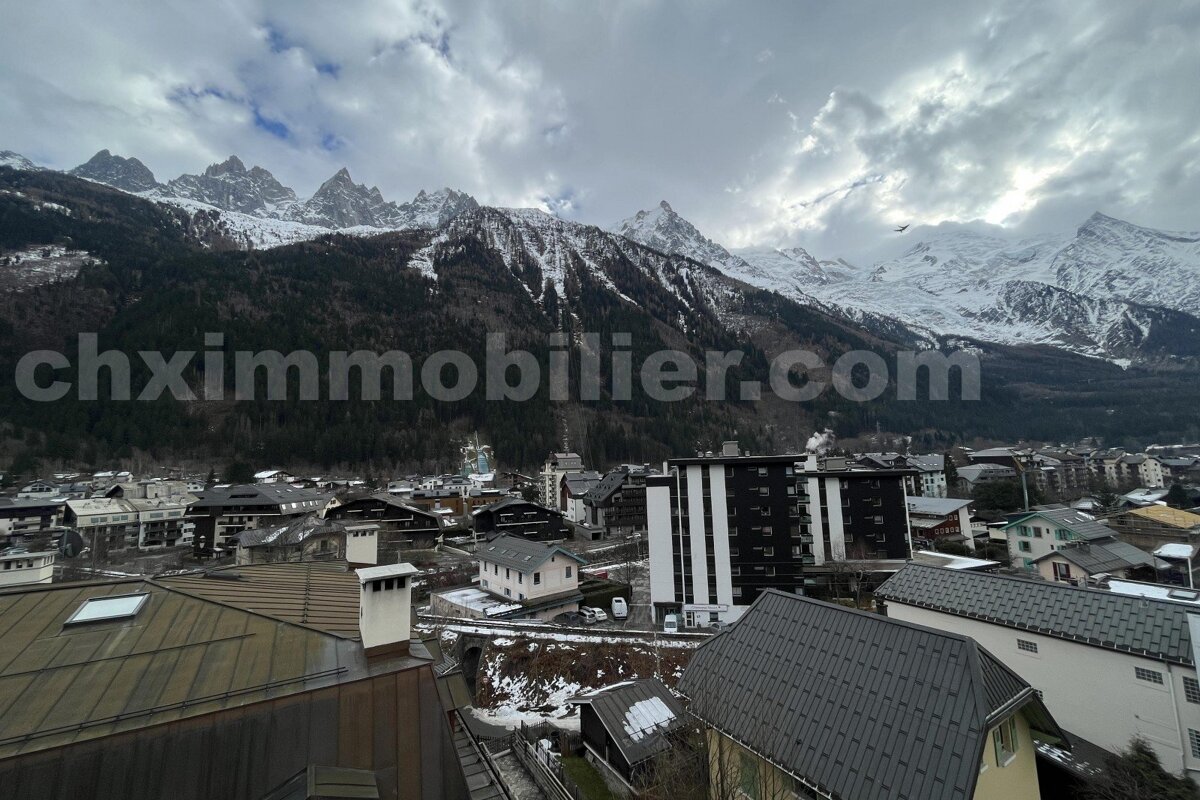 A mountain town nestled beneath majestic, snow-capped peaks under a cloudy sky. Buildings with varying rooflines fill the foreground. 