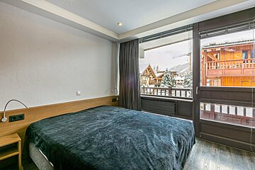 A modern bedroom with a dark bed, wooden headboard, and large window showcasing a snowy mountain village with chalets and pine trees.