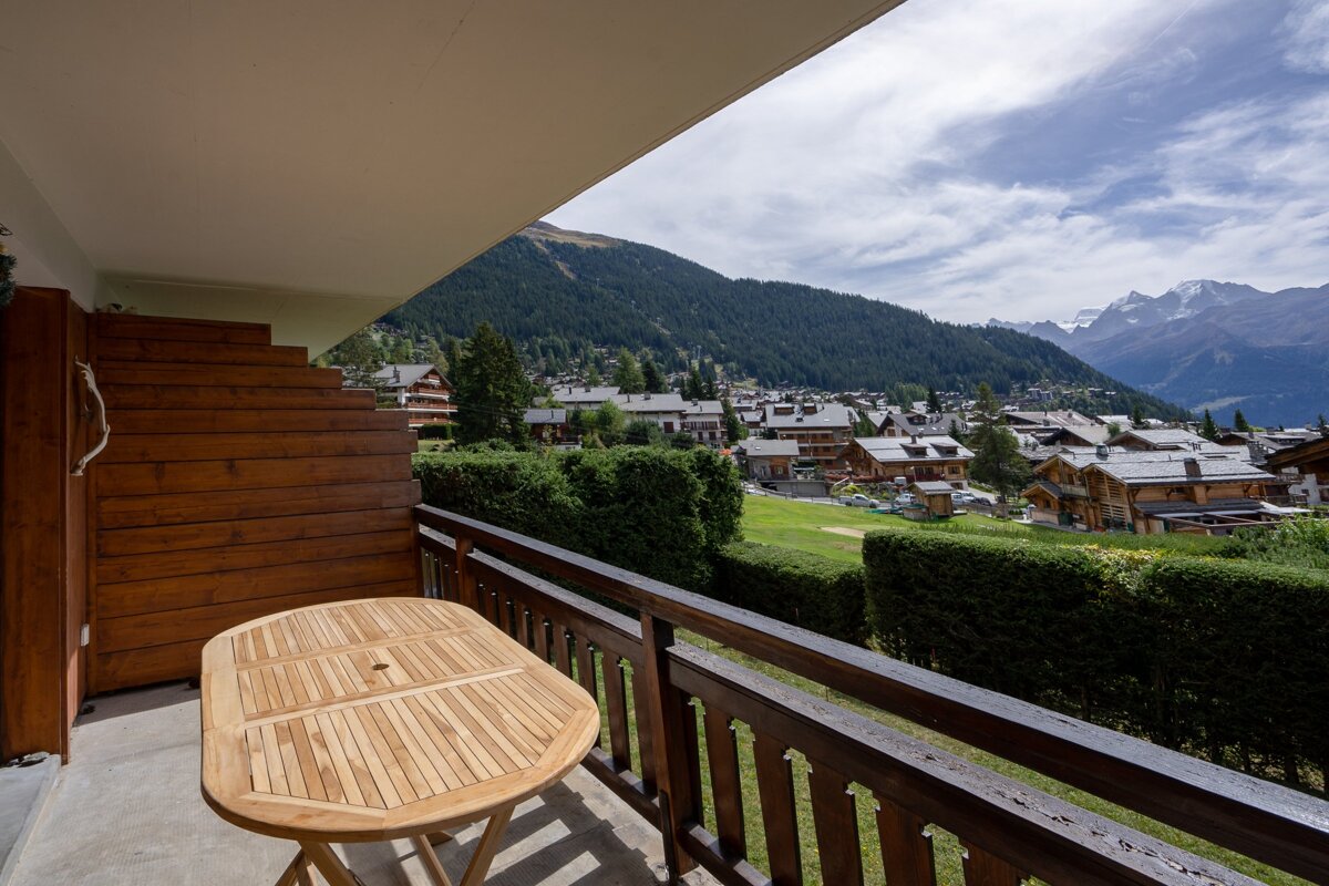 A wooden balcony with a table overlooking a scenic alpine village nestled in the green mountains.