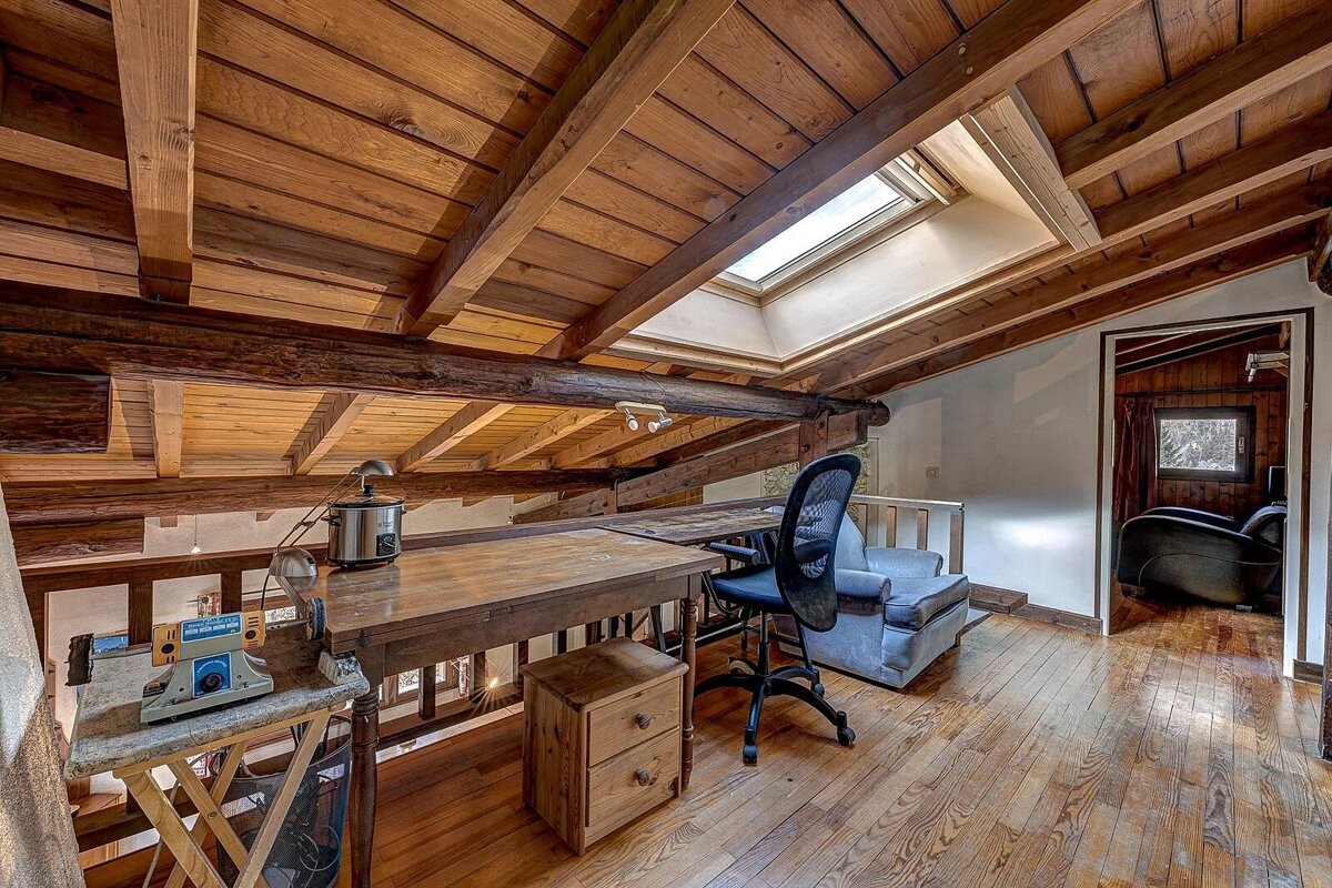 A rustic wooden attic/loft office with a skylight, exposed beams, a desk, office chair, and an armchair.