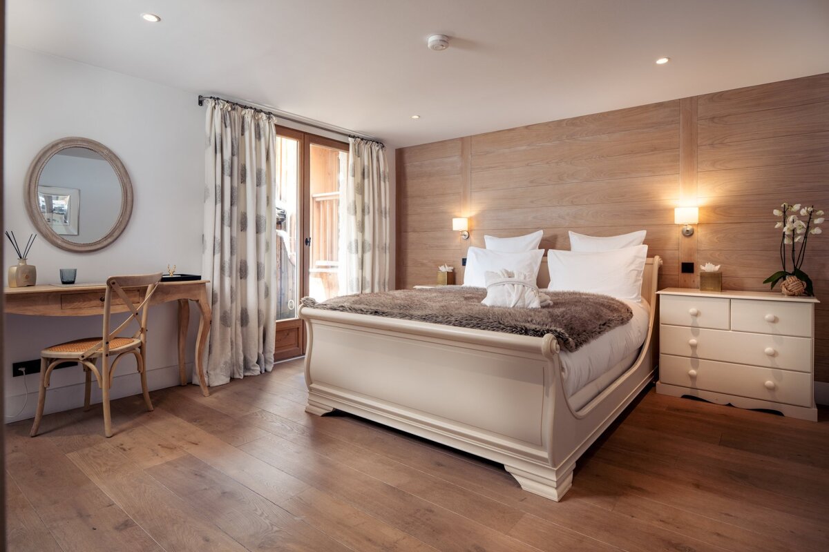 Elegant bedroom featuring a cream sleigh bed, wood-paneled accent wall, writing desk with a round mirror, and warm hardwood floors.