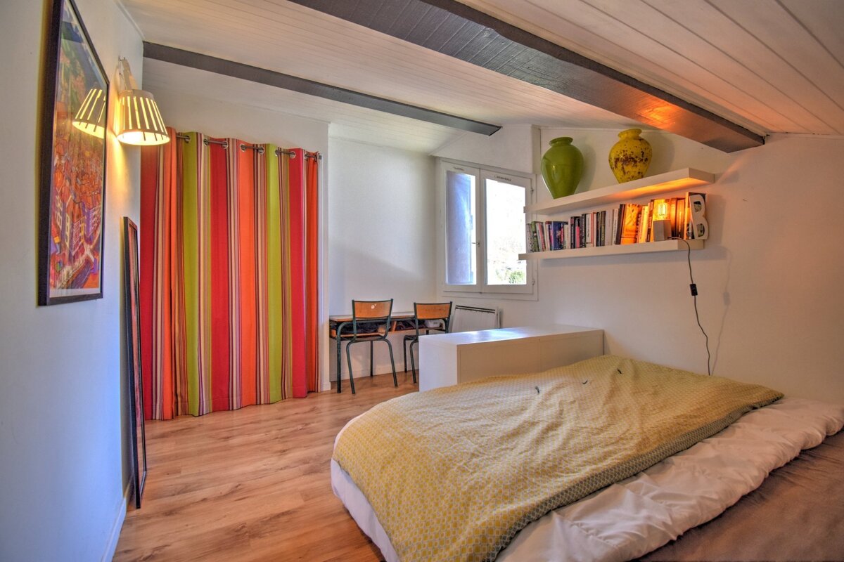 Cozy, bright room with a bed, wooden floor, and vibrant striped curtains. Features exposed beams, white walls, and bookshelves with warm lighting.