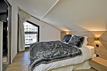 A stylish attic bedroom featuring a plush gray fur blanket, light wood headboard, sloped ceiling, and a window view of urban buildings.