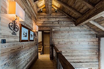 A warm, rustic wooden chalet interior hallway/landing with exposed beams, wall sconces, framed art, and a wooden railing.