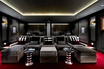 A luxurious home theater features plush gray recliners with ottomans on a red carpet, dark paneled walls, and subtle cove and accent lighting.