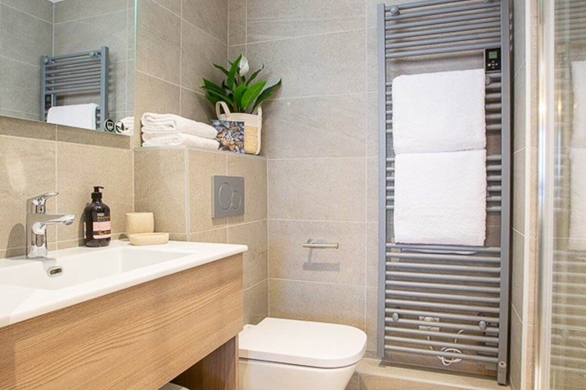 A modern bathroom featuring a wooden vanity, white toilet, and a grey heated towel rack with white towels against grey tiled walls.