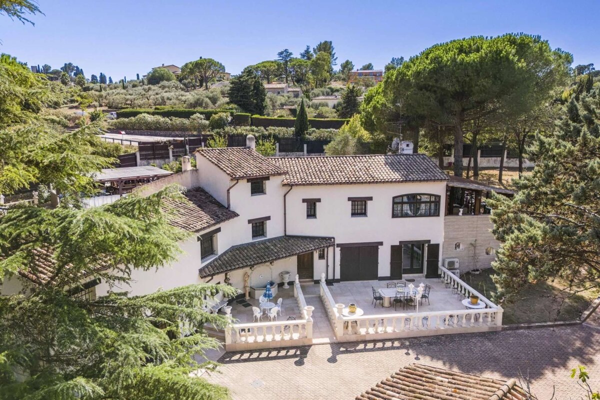 Aerial view of a charming white house with tiled roofs and spacious patios, nestled amidst green hills and abundant trees under a clear blue sky.
