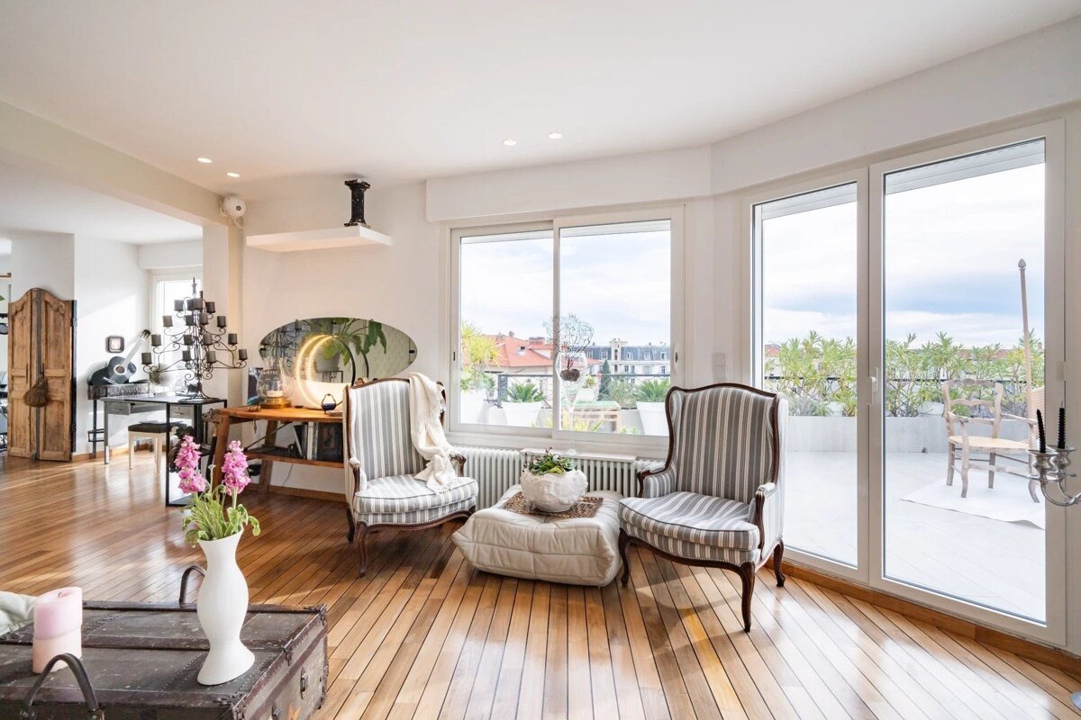 Bright, elegant living room with hardwood floors, two striped armchairs, large windows to a balcony with green plants, and a city view.