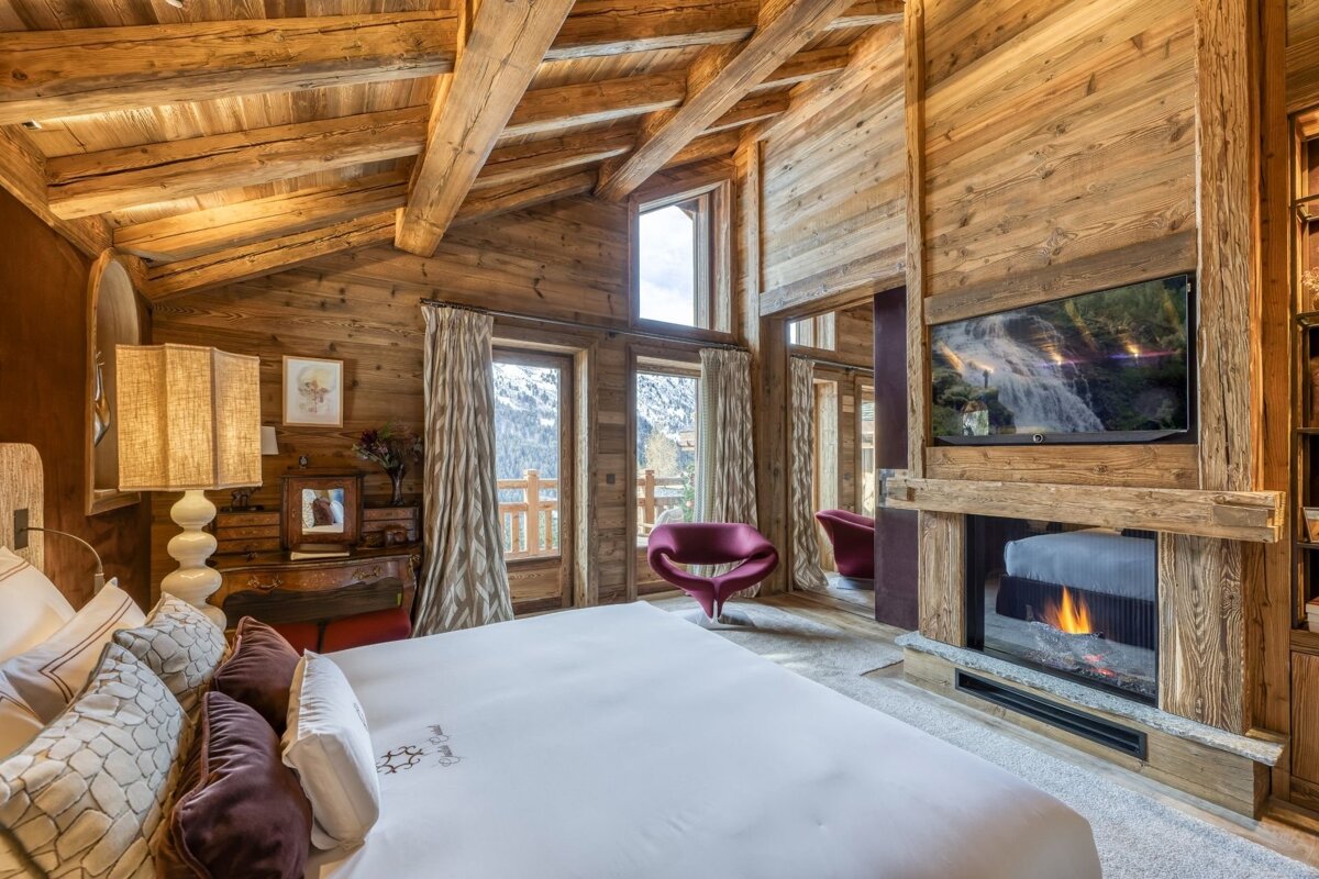 Luxurious chalet bedroom with wood-paneled walls & ceiling, a cozy fireplace, TV, plush bed, and stunning mountain views from large windows.