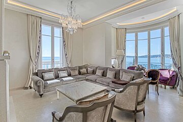 Luxurious living room with a large gray sectional sofa, crystal chandelier, and expansive windows offering a stunning ocean view. Elegant purple chairs.