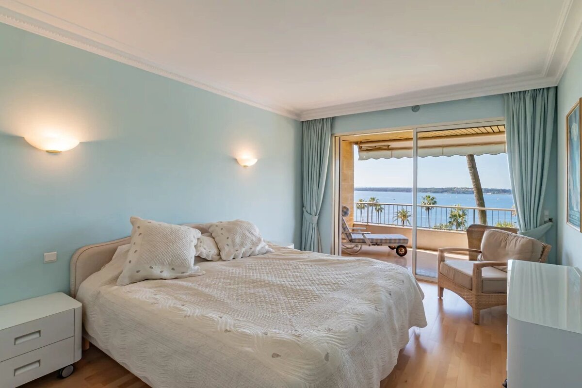 Bright bedroom with light blue walls, a large bed, and sliding glass doors leading to a balcony with a scenic ocean view.