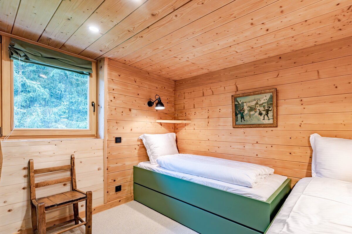 A rustic wooden bedroom features two beds with green bases, a forest-view window, a vintage painting, and a cozy wall lamp.